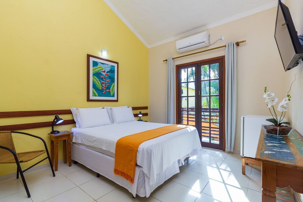 A hotel room with a bed, a desk with a computer, a chair, a painting on the wall, and a door leading to an outdoor area.