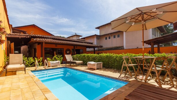 Una piscina con sillones de descanso y una sombrilla, rodeada por un patio con baldosas y un seto.