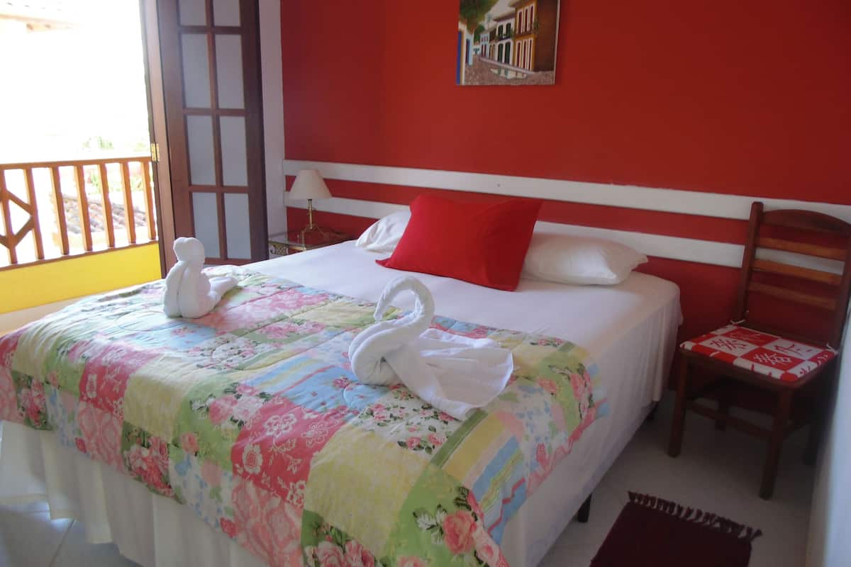 A hotel room with a bed, a red wall, a wooden chair, and a window with a view.