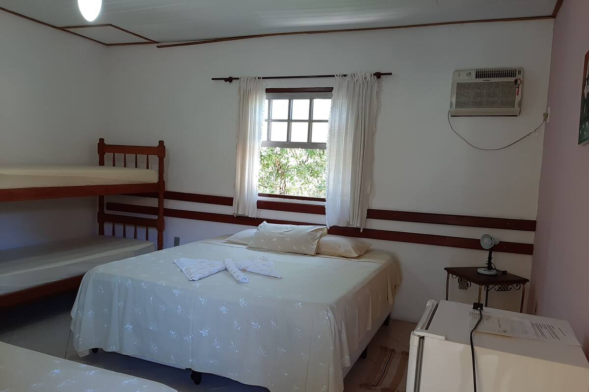 A room with a bunk bed, a single bed, a window with curtains, a wall-mounted air conditioner, and a desk with a lamp.