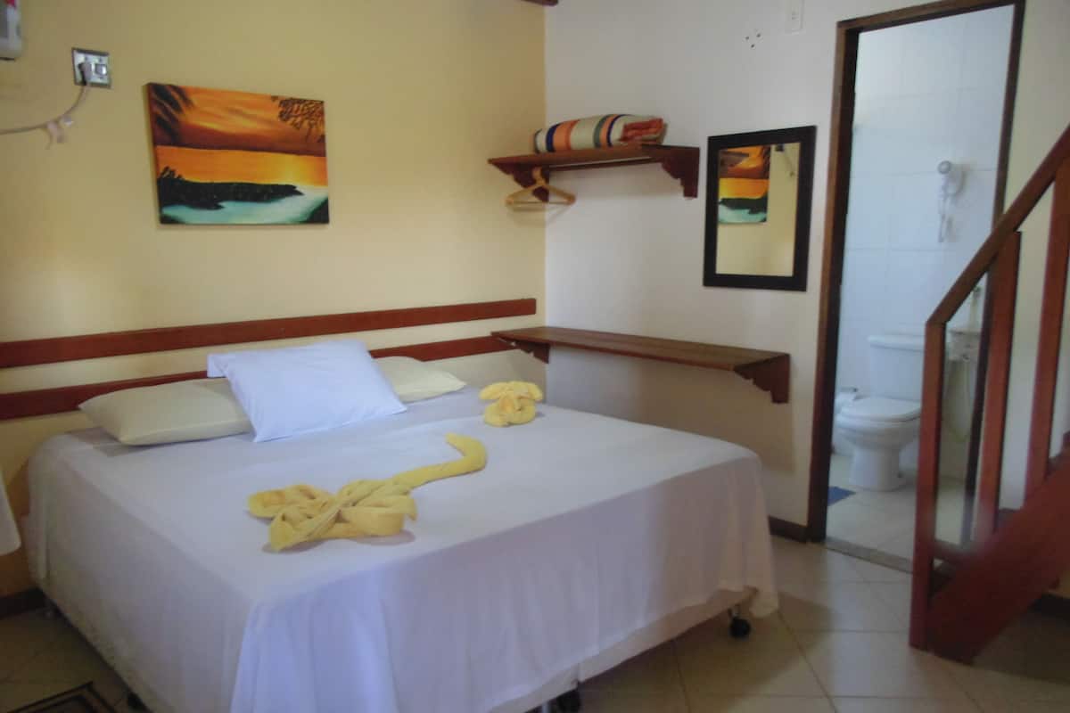 A hotel room with a bed, pillows, a bedside table, a painting on the wall, a shelf with towels, and a mirror.