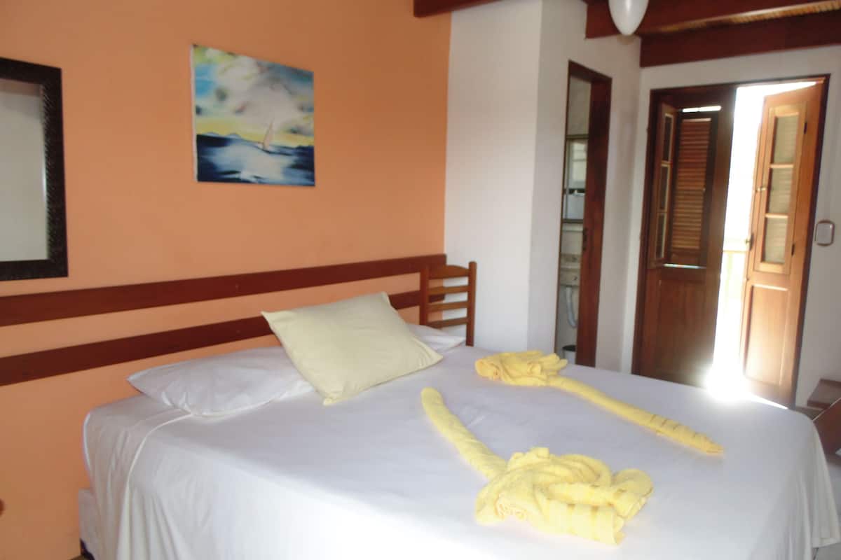 A neatly made bed with a white bedspread and pillows, a painting of a sailboat on the wall, and a wooden door leading to another room.