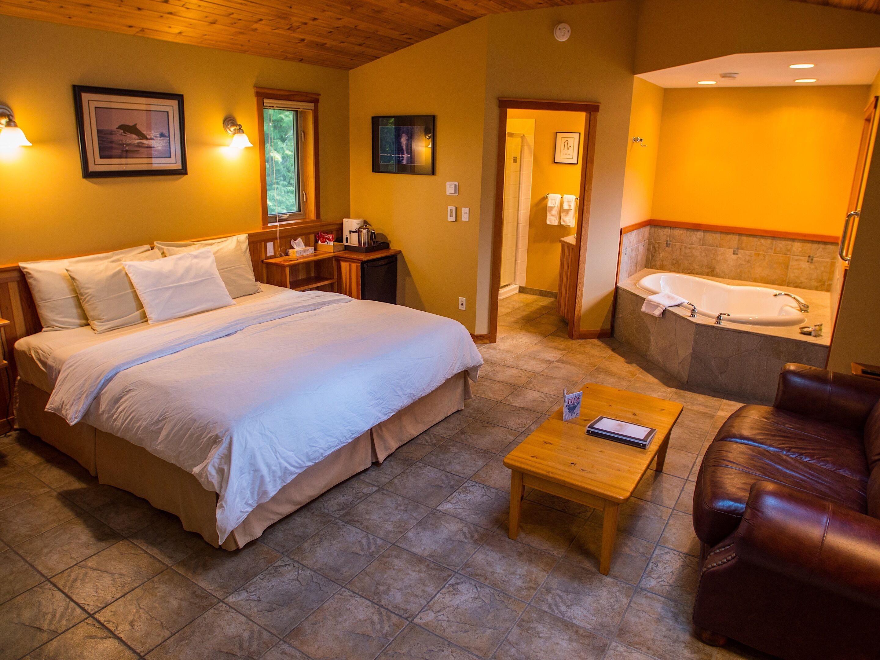 Une chambre d’hôtel avec un grand lit, une table basse en bois, un canapé en cuir, une salle de bain avec baignoire et un petit bureau avec une lampe.
