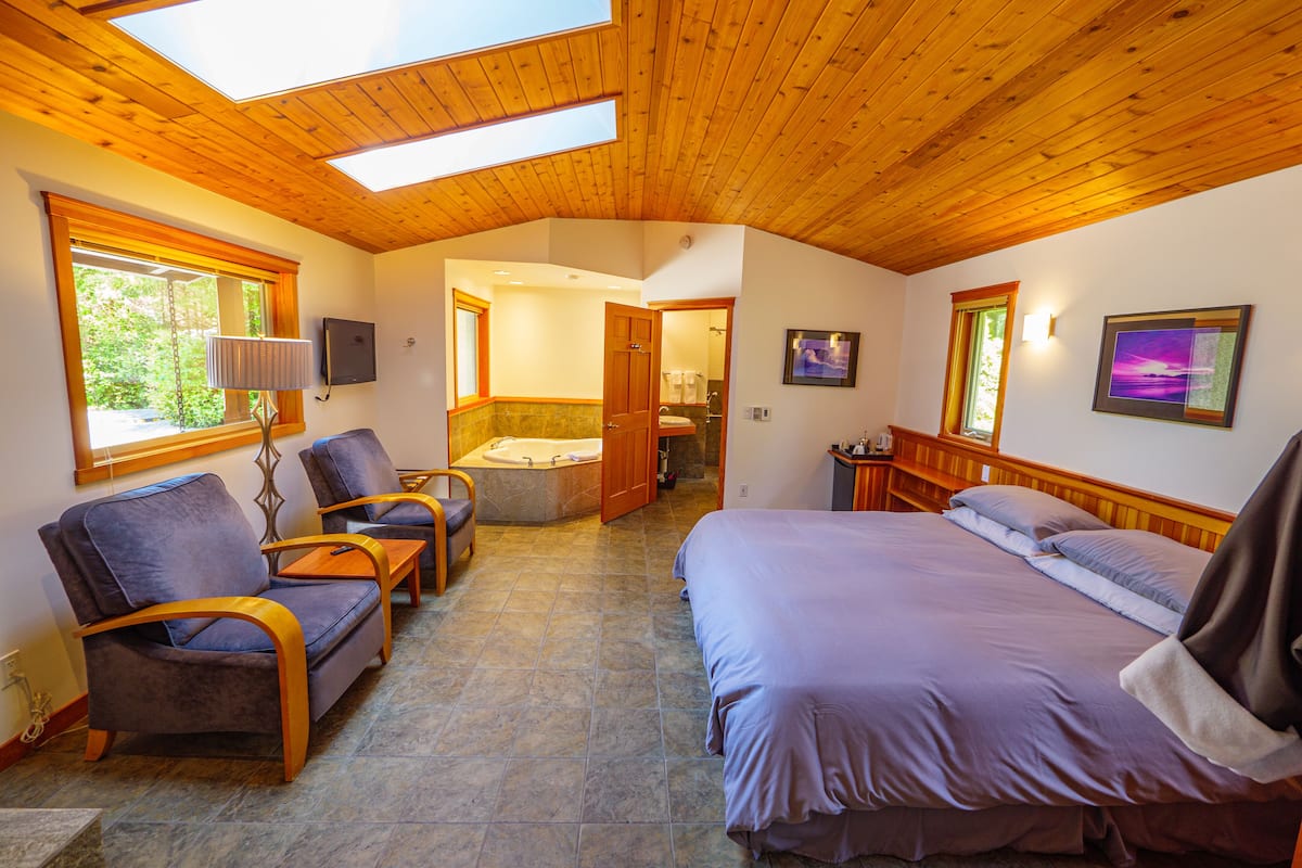 A room with a bed, two armchairs, a small table, a bathtub, and a window with a view of greenery.