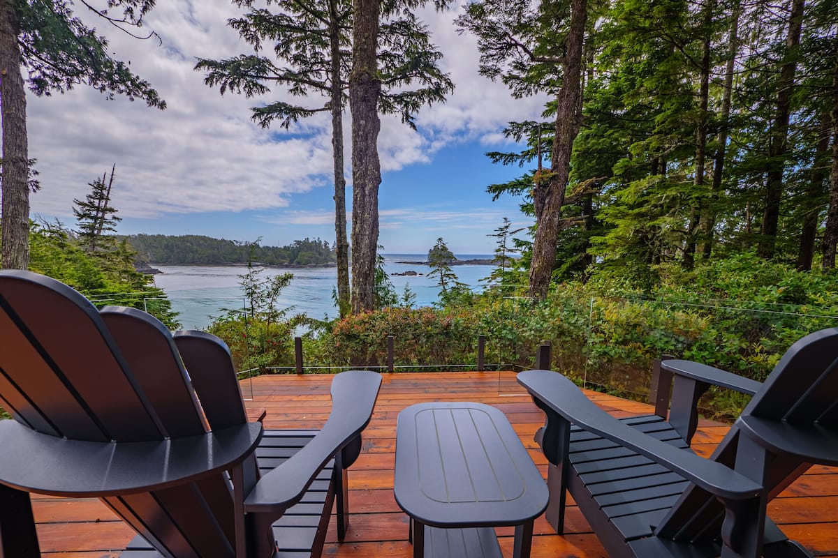 A deck with Adirondack chairs and a picnic table overlooking a forested area and a body of water.