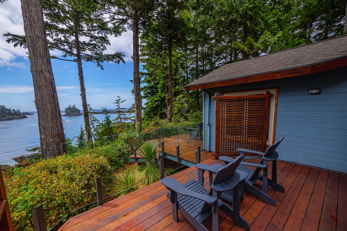 A wooden deck with two Adirondack chairs, overlooking a body of water and surrounded by trees.