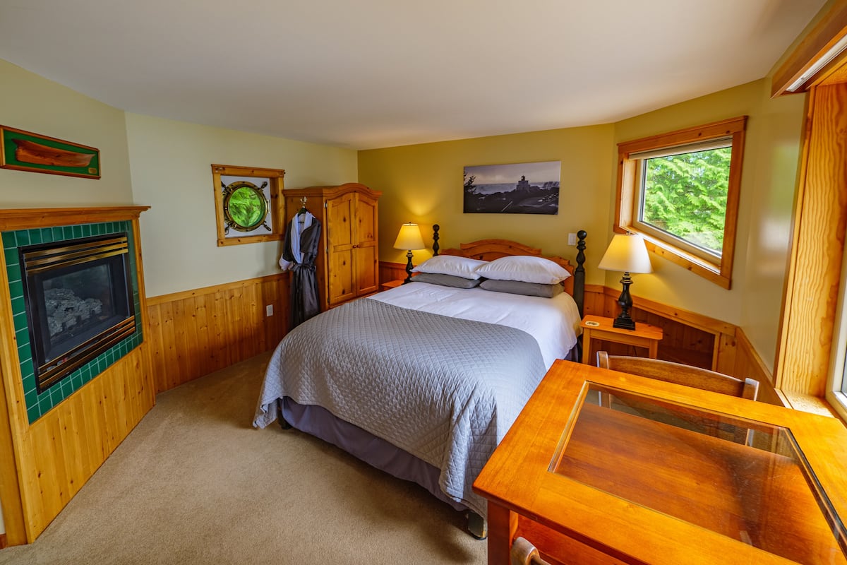 A bedroom with a bed, a wooden wardrobe, a fireplace, a painting, and a window with a view.