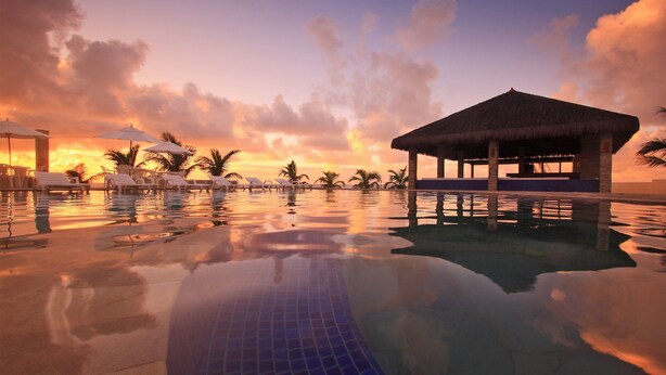 A serene pool area with lounge chairs and umbrellas, a thatched-roof structure, and a sunset view. - Village Porto de Galinhas (Ipojuca)