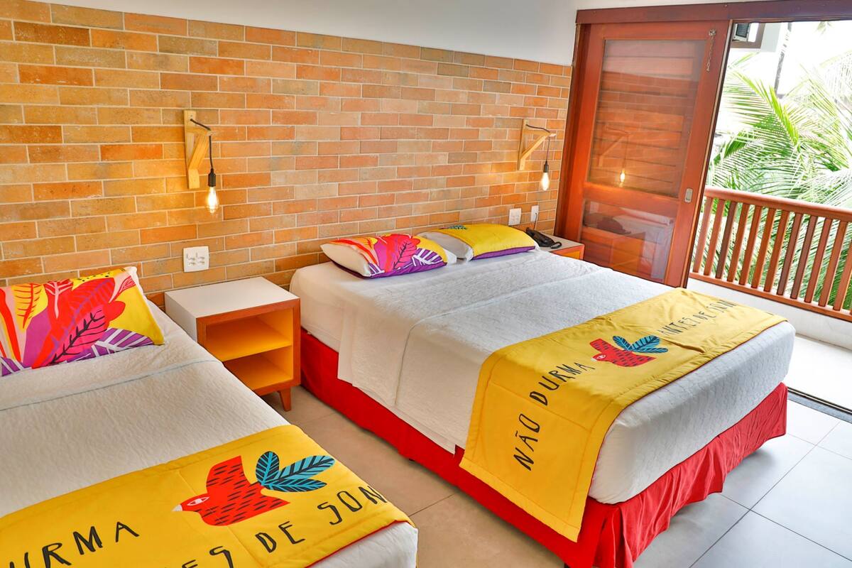 Ein Hotelzimmer mit zwei Betten, jedes mit einem farbenfrohen Kopfkissen und einer gelben Bettdecke mit Aufdruck. An der Wand sind Leuchten befestigt und es gibt einen Blick ins Grüne.