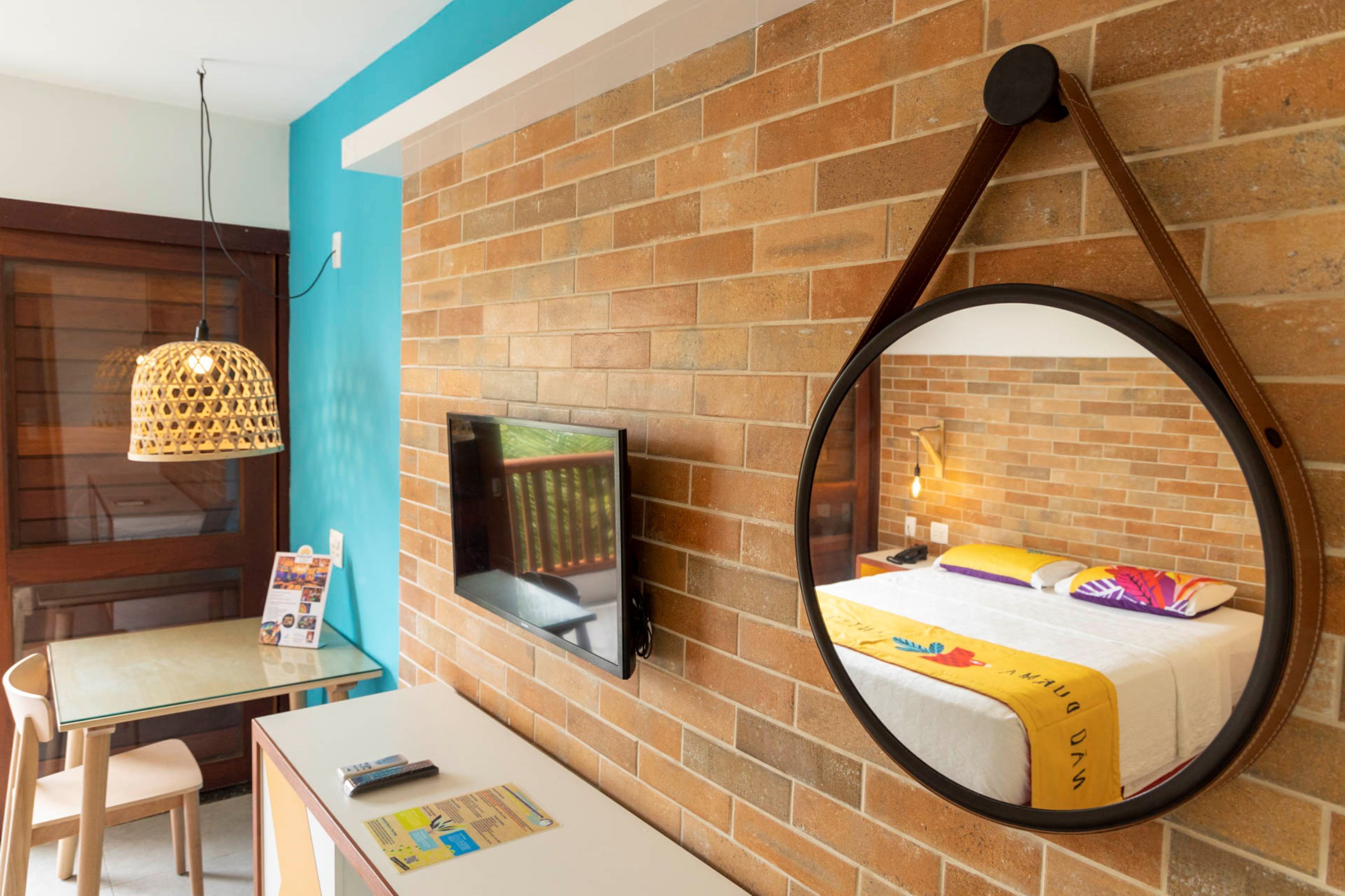 A hotel room with a brick wall, a round mirror, a bed with a colorful blanket, a TV mounted on the wall, a table with chairs, and a hanging light fixture.