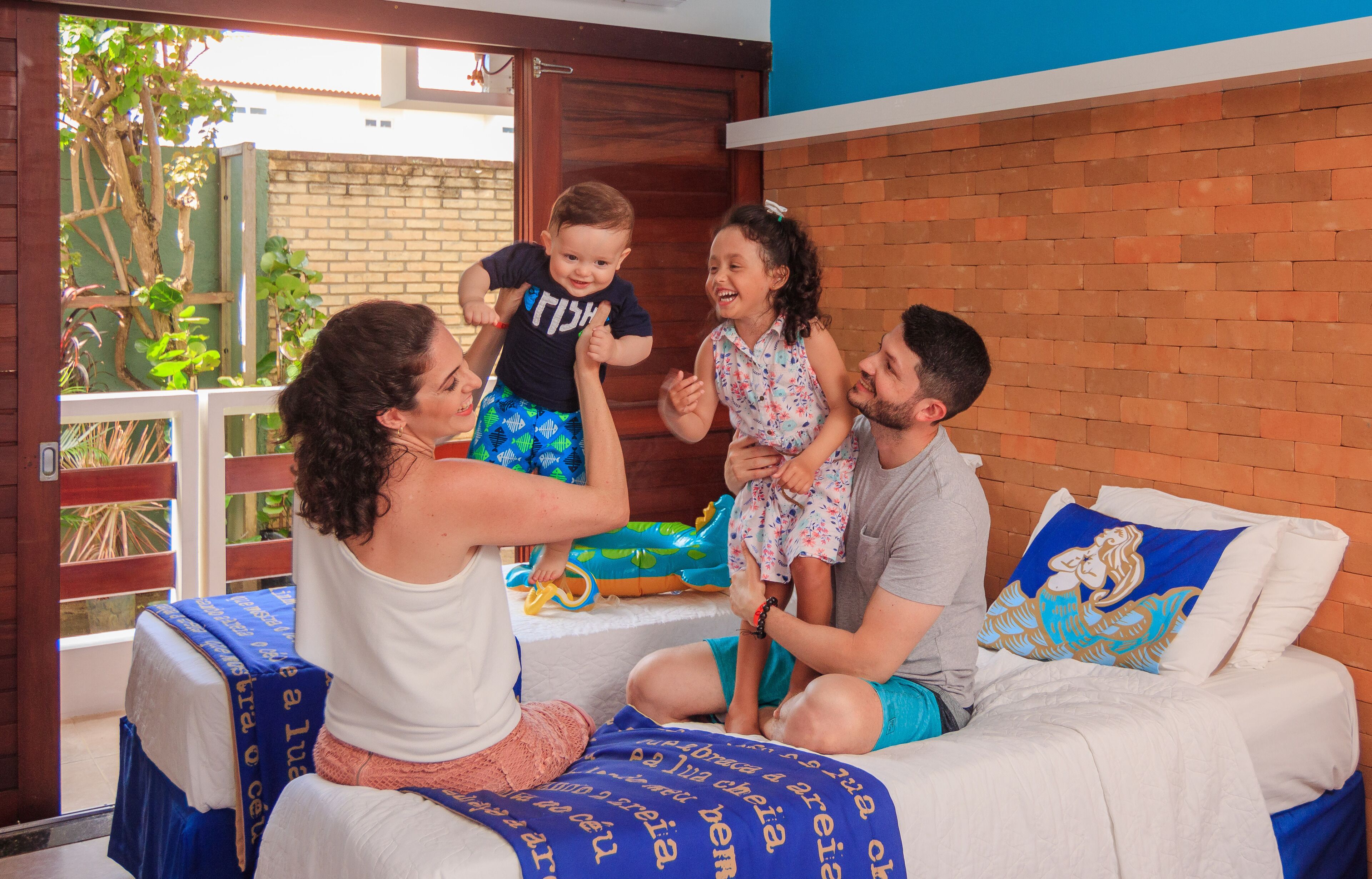 Una famiglia di quattro persone su un letto con una coperta a motivi blu e bianchi, con un muro di mattoni e una porta di legno sullo sfondo.