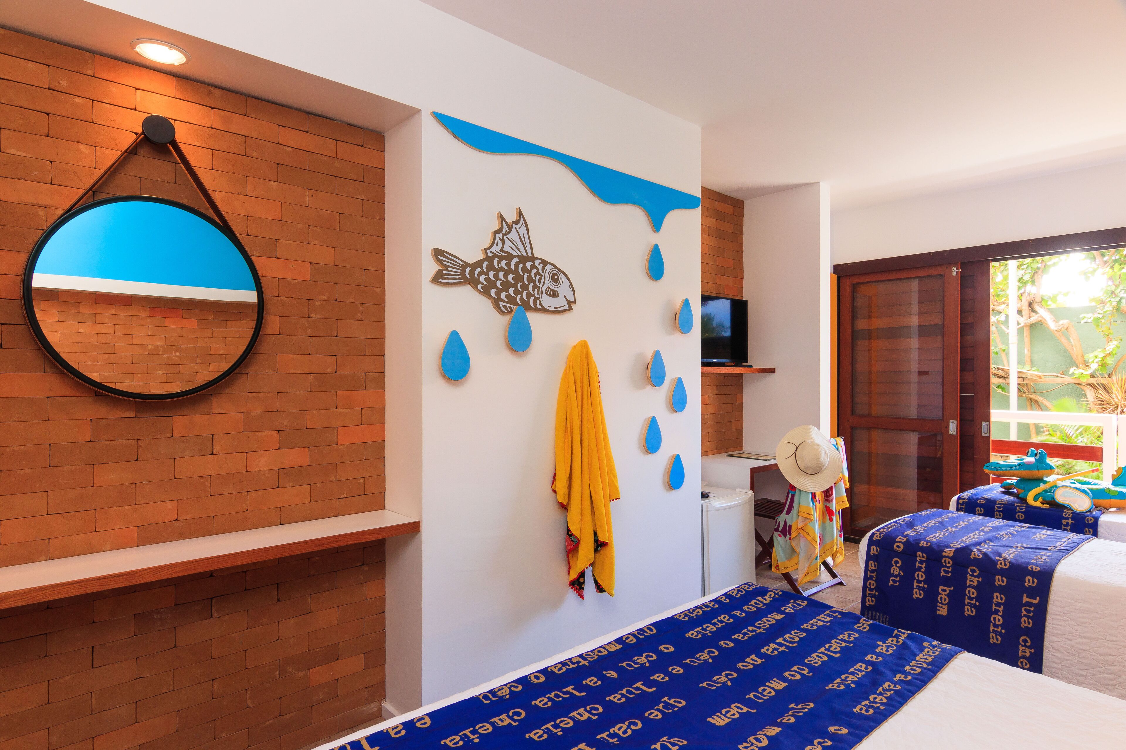 Una habitación de hotel con pared de ladrillo, un espejo redondo, una decoración de pez, una toalla amarilla, un sombrero y una cama con cubrecama de estampado azul.