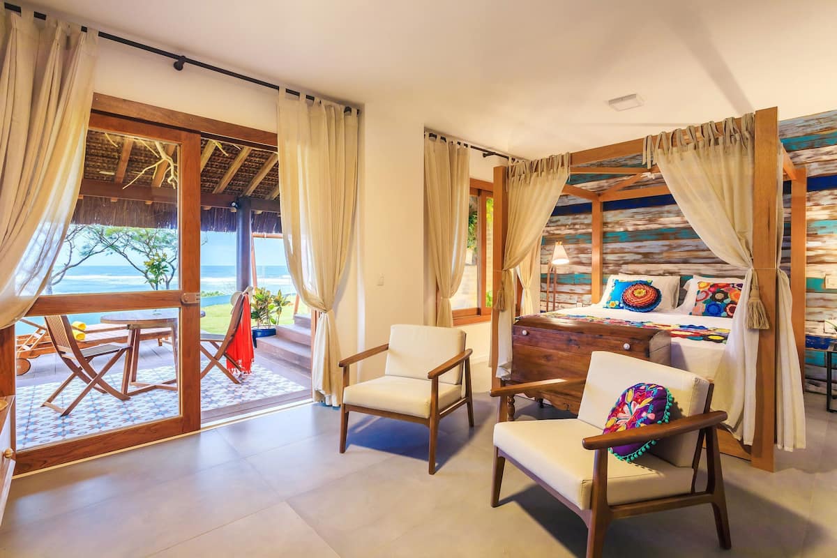 Ein Zimmer mit Holzboden, einem Bett mit bunten Kissen und Blick auf das Meer durch große Glasfenster.