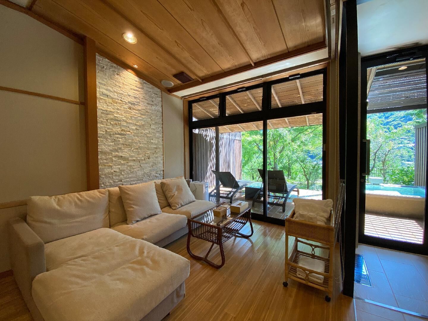 Una sala de estar moderna con un sofá, una mesita y una silla de mimbre. Se ve un ventanal con vistas a la vegetación.