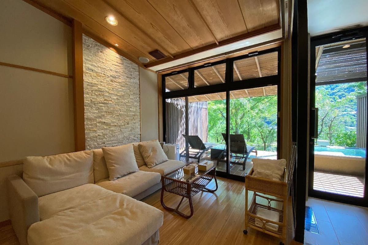 Una sala de estar moderna con un sofá, una mesita y una silla de mimbre. Se ve un ventanal con vistas a la vegetación.