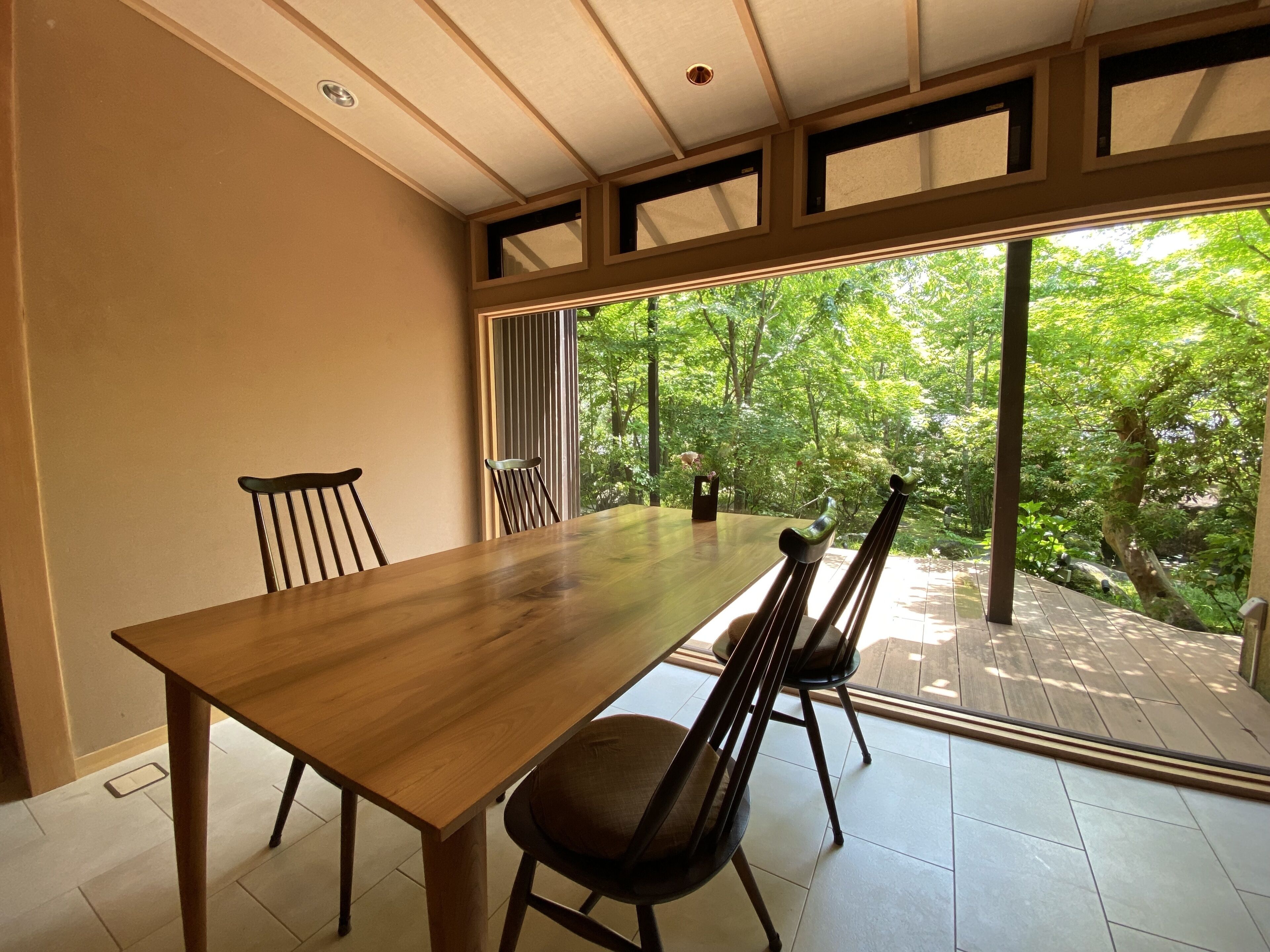 A dining area with a wooden table and chairs, large windows with a view of greenery outside.
