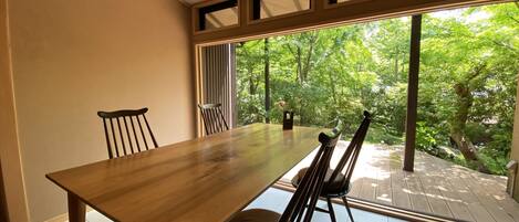 A dining area with a wooden table and chairs, large windows with a view of greenery outside.