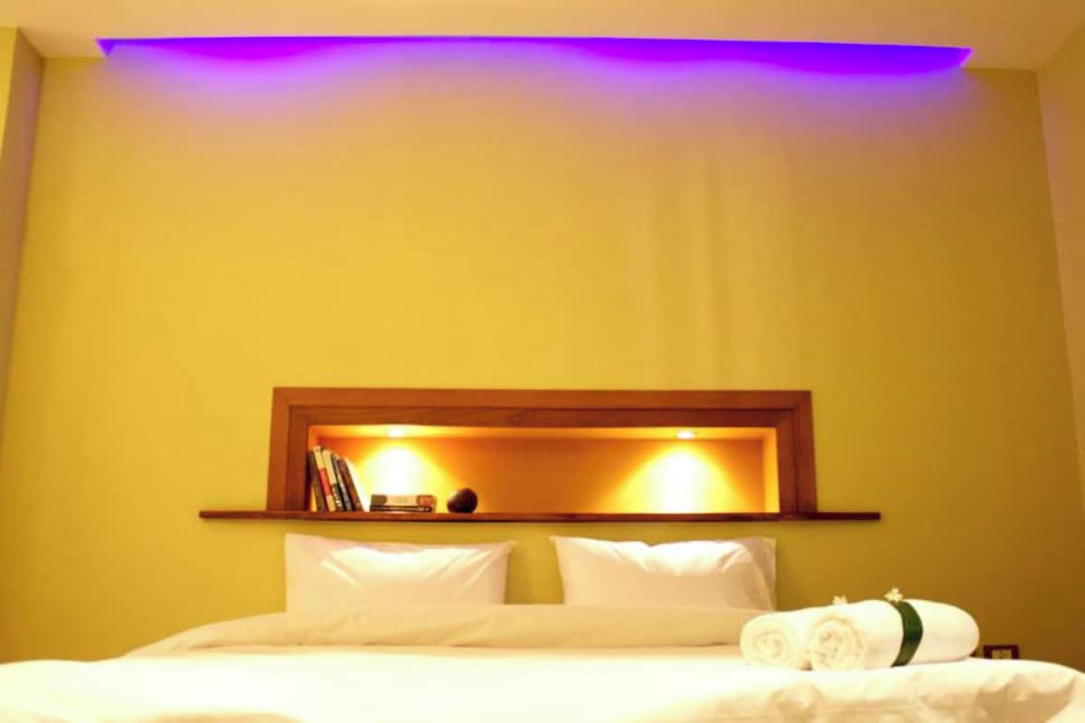 Quarto de hotel com cama, almofadas, mesa de cabeceira com livros e prateleira na parede.