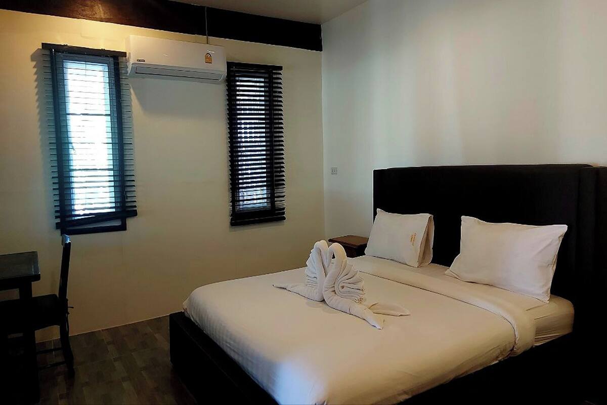 Una habitación de hotel con cama, dos ventanas con persianas, una mesita de noche y una silla.
