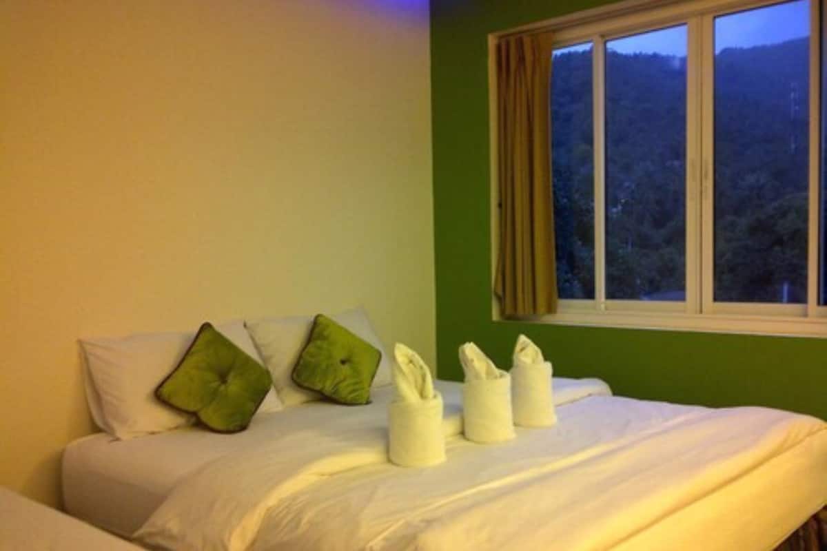 Una habitación de hotel con una cama, almohadillas verdes, ropa de cama blanca y una ventana con vistas a un bosque.