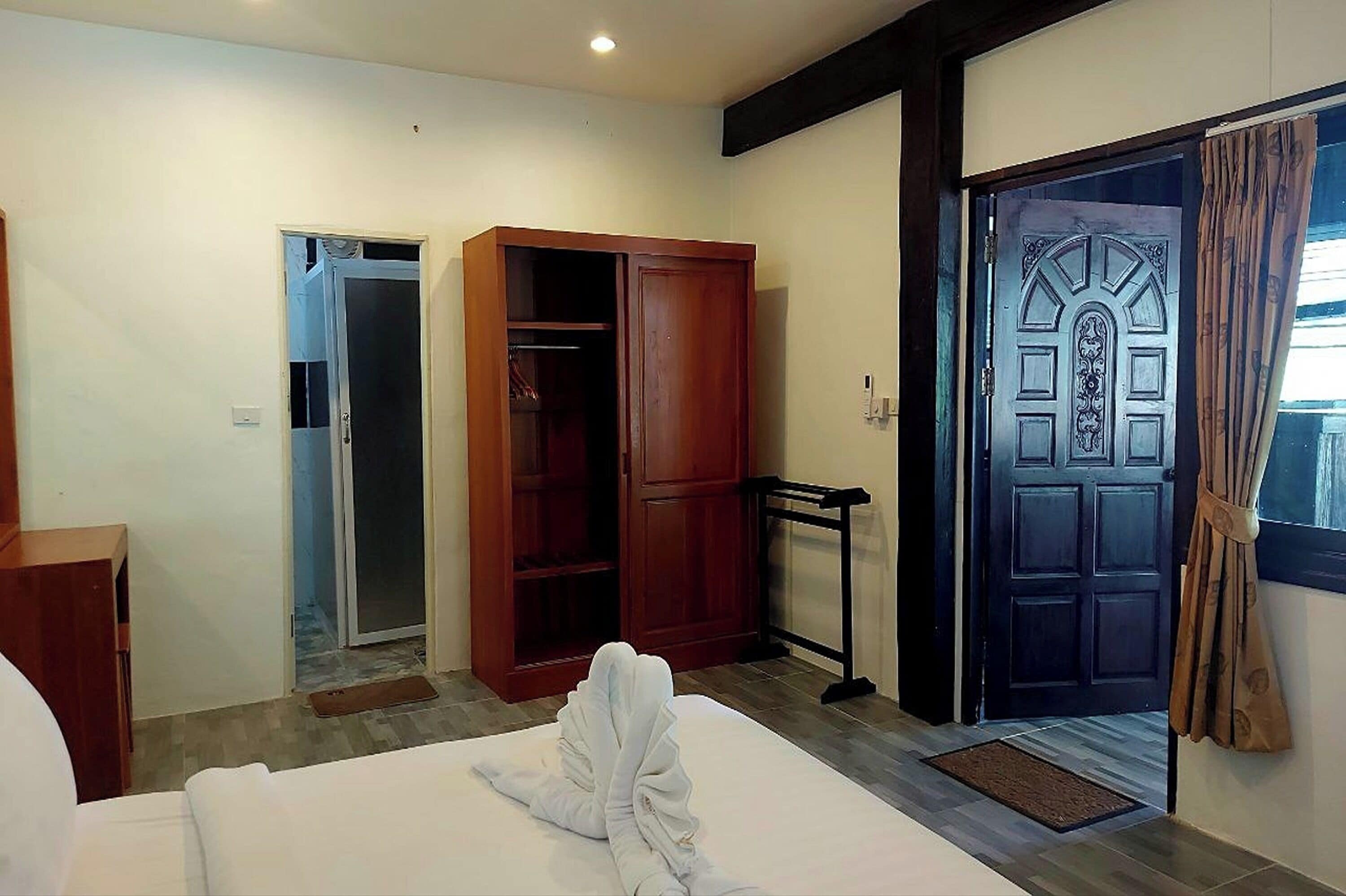 A hotel room with a bed, a wooden wardrobe, a door with a decorative design, and a small window with curtains.