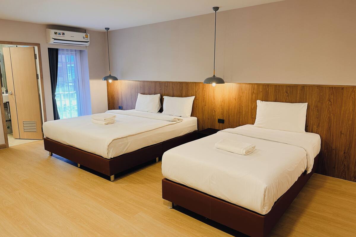 A hotel room with two beds, wooden headboards, a large window with curtains, an air conditioning unit, and a bedside table with a lamp.
