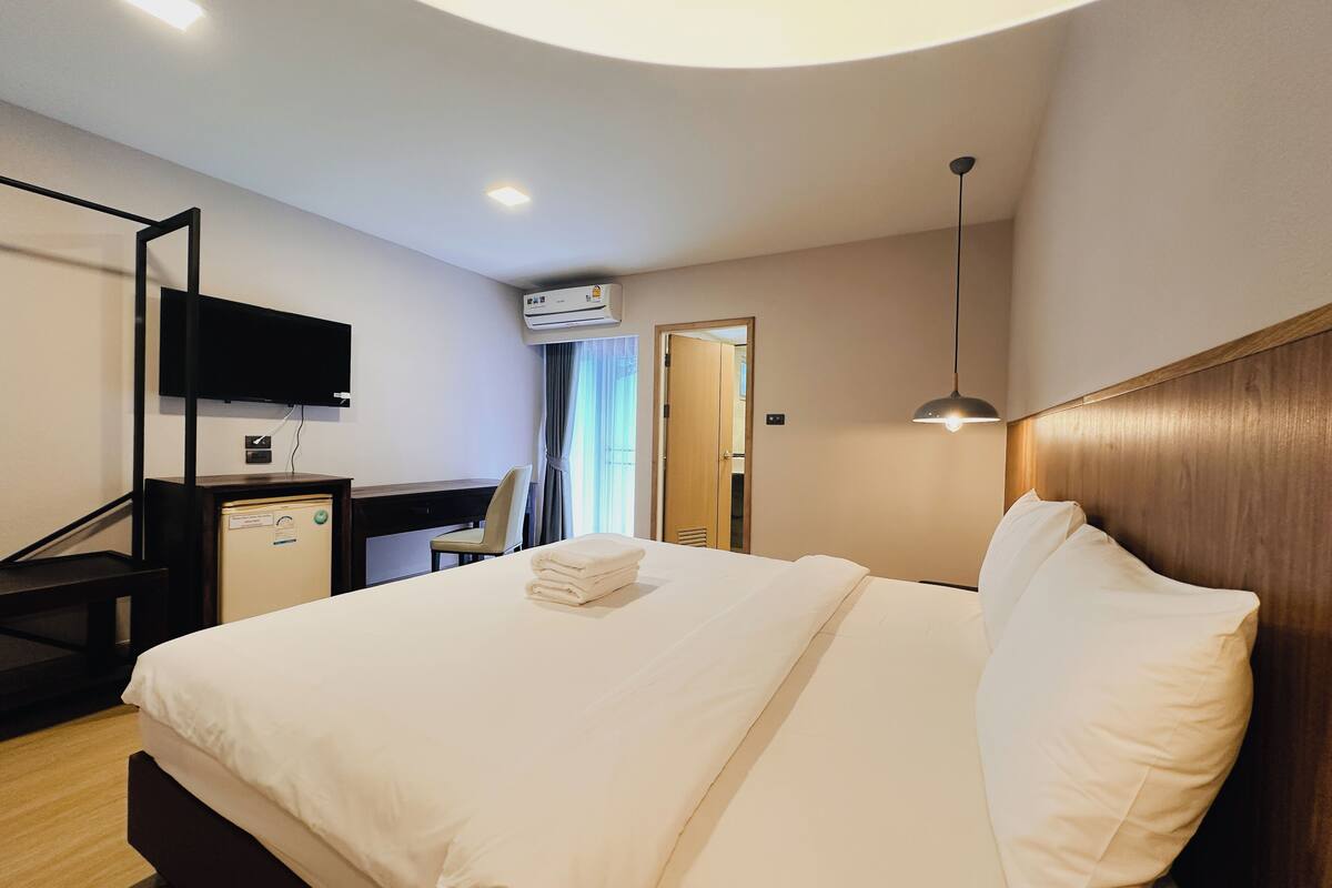 A hotel room with a bed, a desk, a chair, a TV mounted on the wall, and an air conditioning unit.