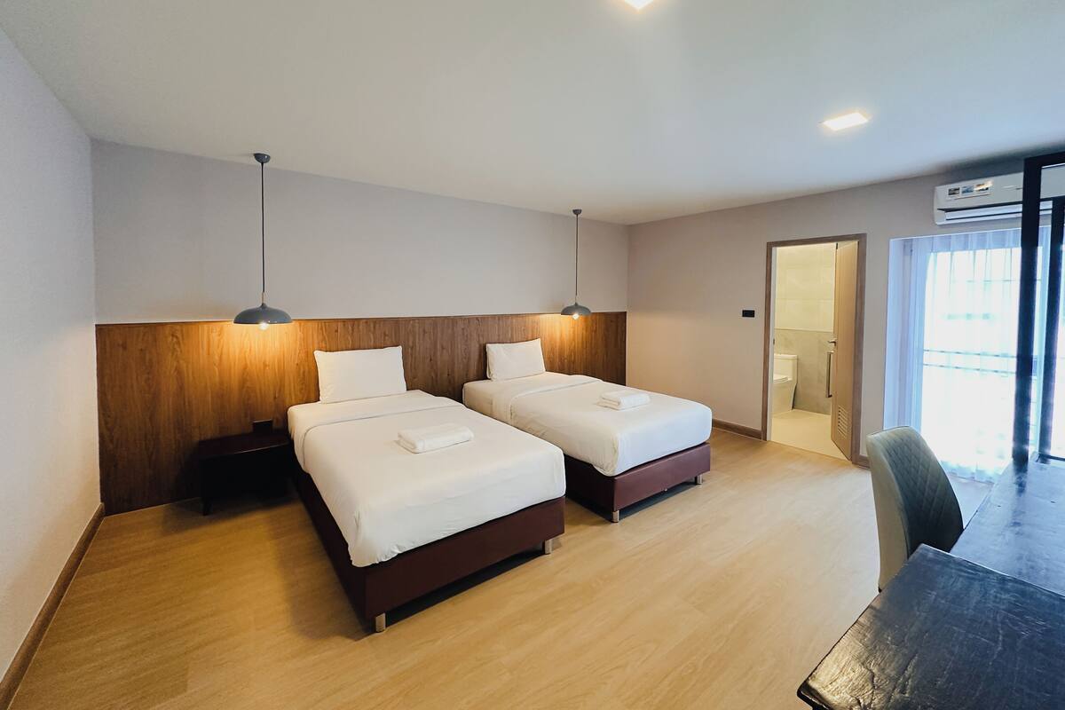 A hotel room with two beds, wooden headboard, modern pendant lights, and a view of the outside through a large window.
