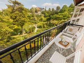 A balcony with a view of a lake, surrounded by greenery and a building with a traditional roof.