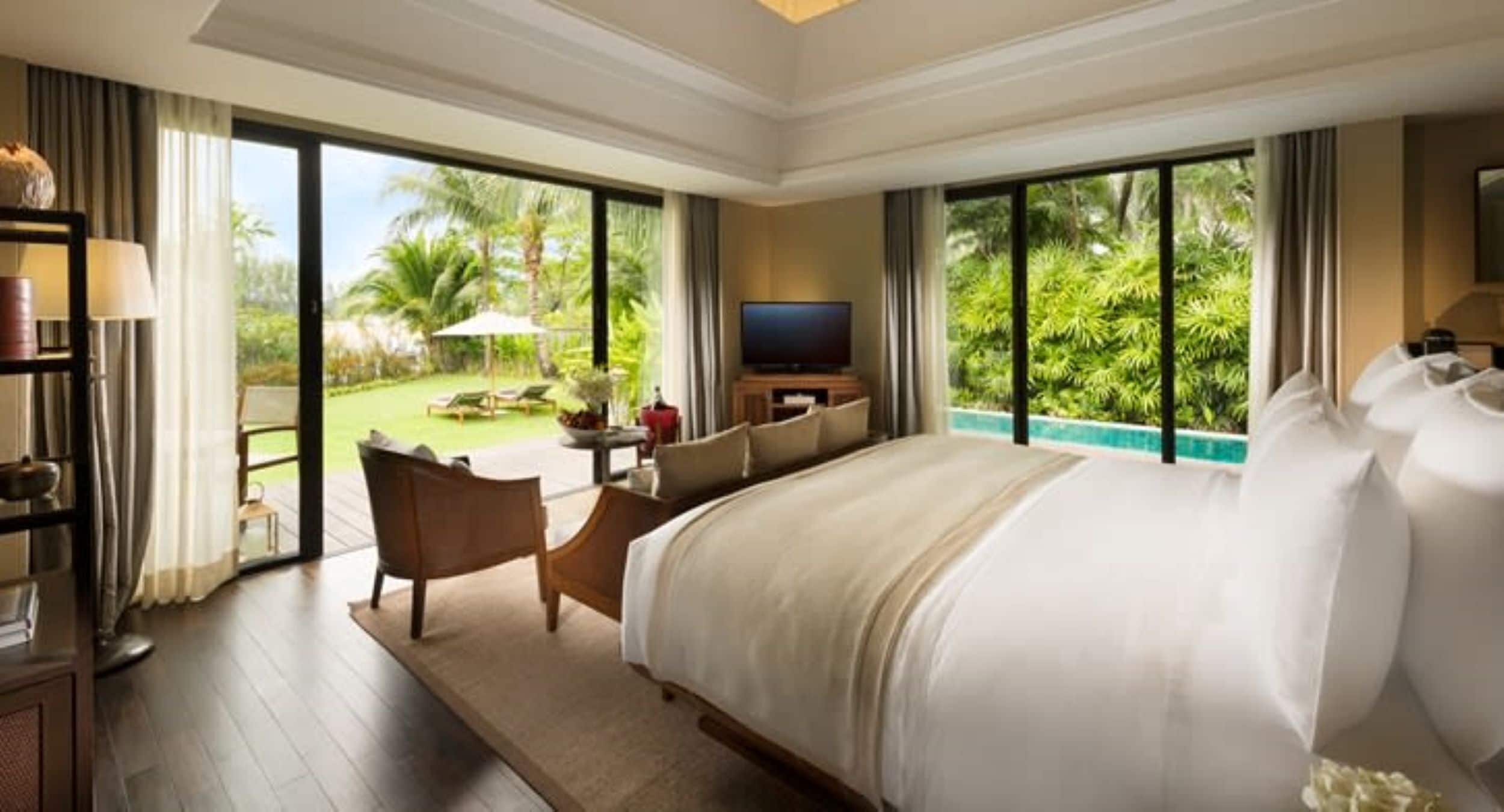 A spacious hotel room with a large bed, a television, and a view of a lush garden and pool.