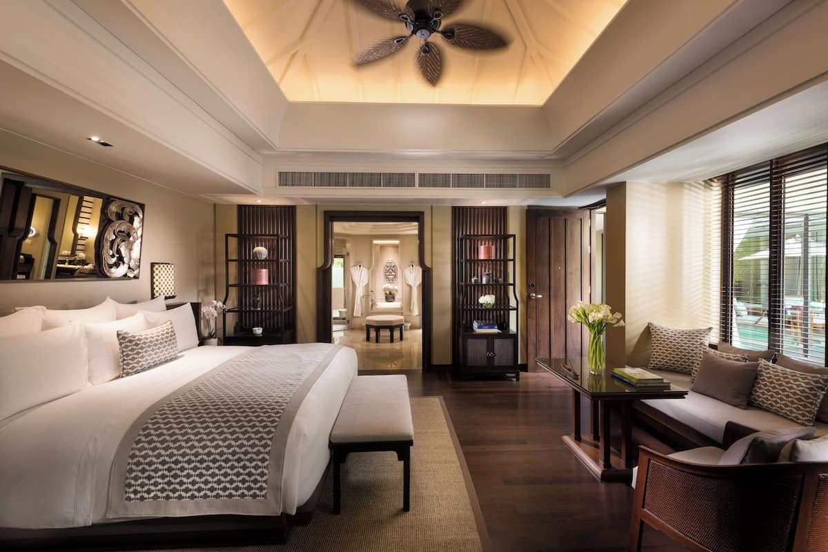A spacious bedroom with a large bed, a sitting area with a sofa and armchairs, a ceiling fan, and a view of the outdoors through large windows.