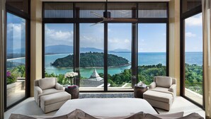 Amplia habitación con ventanas grandes que ofrecen vista a una isla tropical y al mar. Hay dos sillones y una cama con colcha blanca.