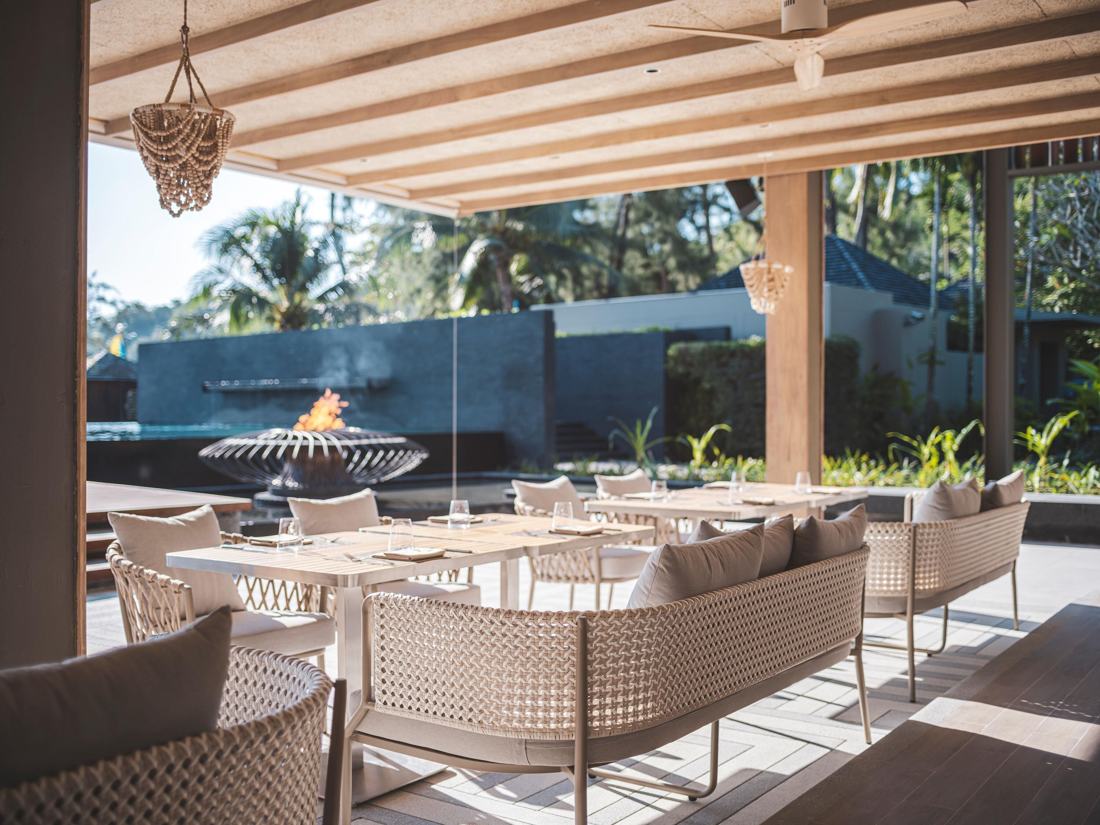 An outdoor dining area with wicker furniture, a fire feature, and a view of a pool and greenery.