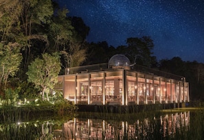 A modern glass building with a dome structure, surrounded by trees and a body of water, under a starry night sky.