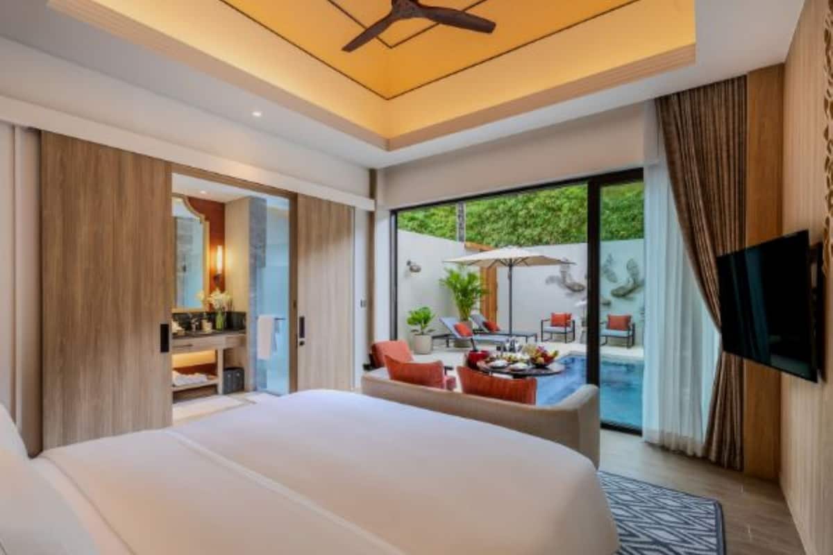 A modern hotel room with a large bed, a TV, a balcony with outdoor seating, and a view of greenery.