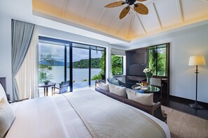 A hotel room with a large bed, a sitting area with a table and chairs, a television, and a view of a lake and hills. - Anantara Layan Phuket Resort (Choeng Thale)