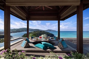 A covered outdoor seating area with a table set for two, overlooking a scenic view of a lake and mountains. - Anantara Layan Phuket Resort (Choeng Thale)