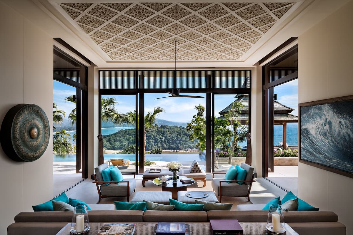 A spacious living area with a large window offering a view of the sea and mountains. There is a sofa set with teal cushions, a coffee table, and a painting on the wall.