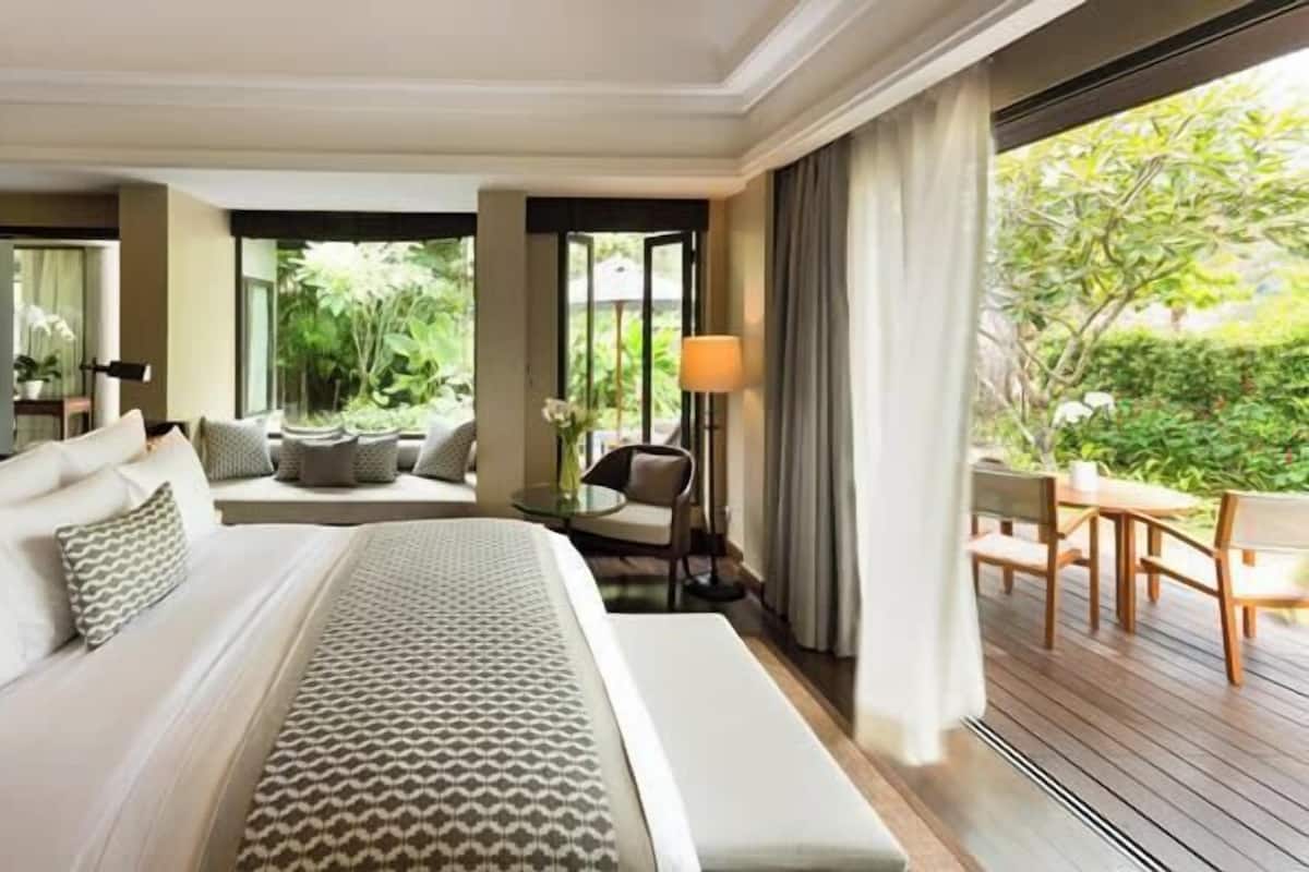 A spacious bedroom with a large bed, a seating area with a sofa and chairs, a small table with a lamp, and a view of greenery outside.