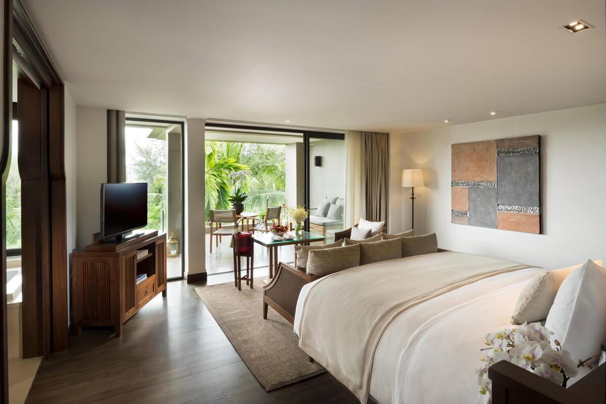 A modern hotel room with a large bed, a television on a wooden stand, a desk with a chair, and a balcony with a view of greenery.