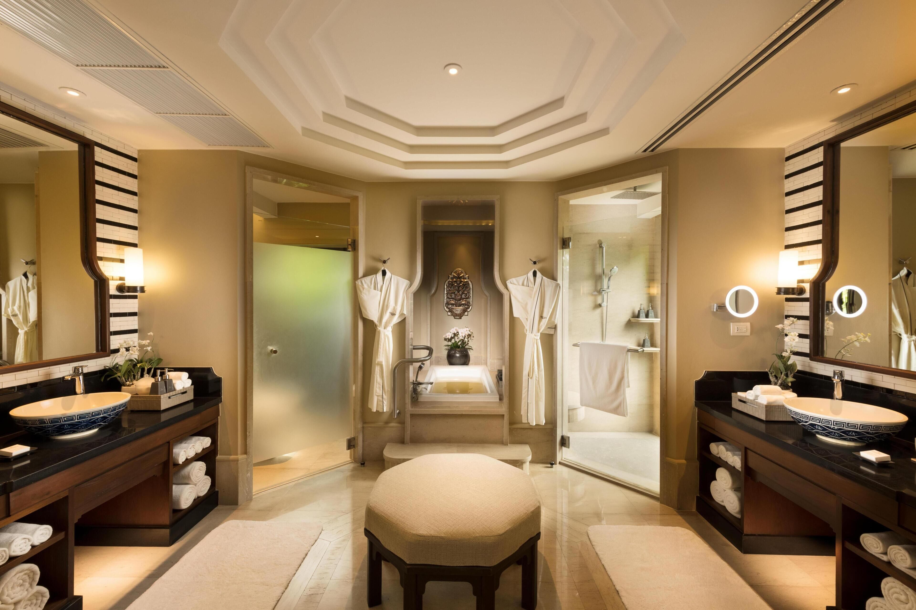 A luxurious bathroom with two large sinks, a bench, and a mirror with lights.