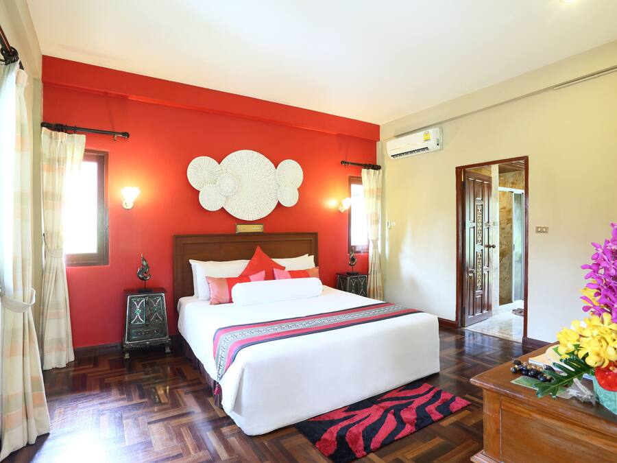 A hotel room with a large bed, a red accent wall, a wooden floor, a nightstand, a mirror, and a floral arrangement.