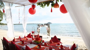 Montaje para comer frente al mar, con una mesa dispuesta para dos, manteles rojos y blancos, servilletas rojas y un centro de mesa floral.