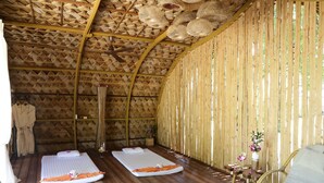 Una habitación con techo y paredes de madera, dos mesas de masaje con toallas, una silla de mimbre y una pequeña mesa con un jarrón de flores.