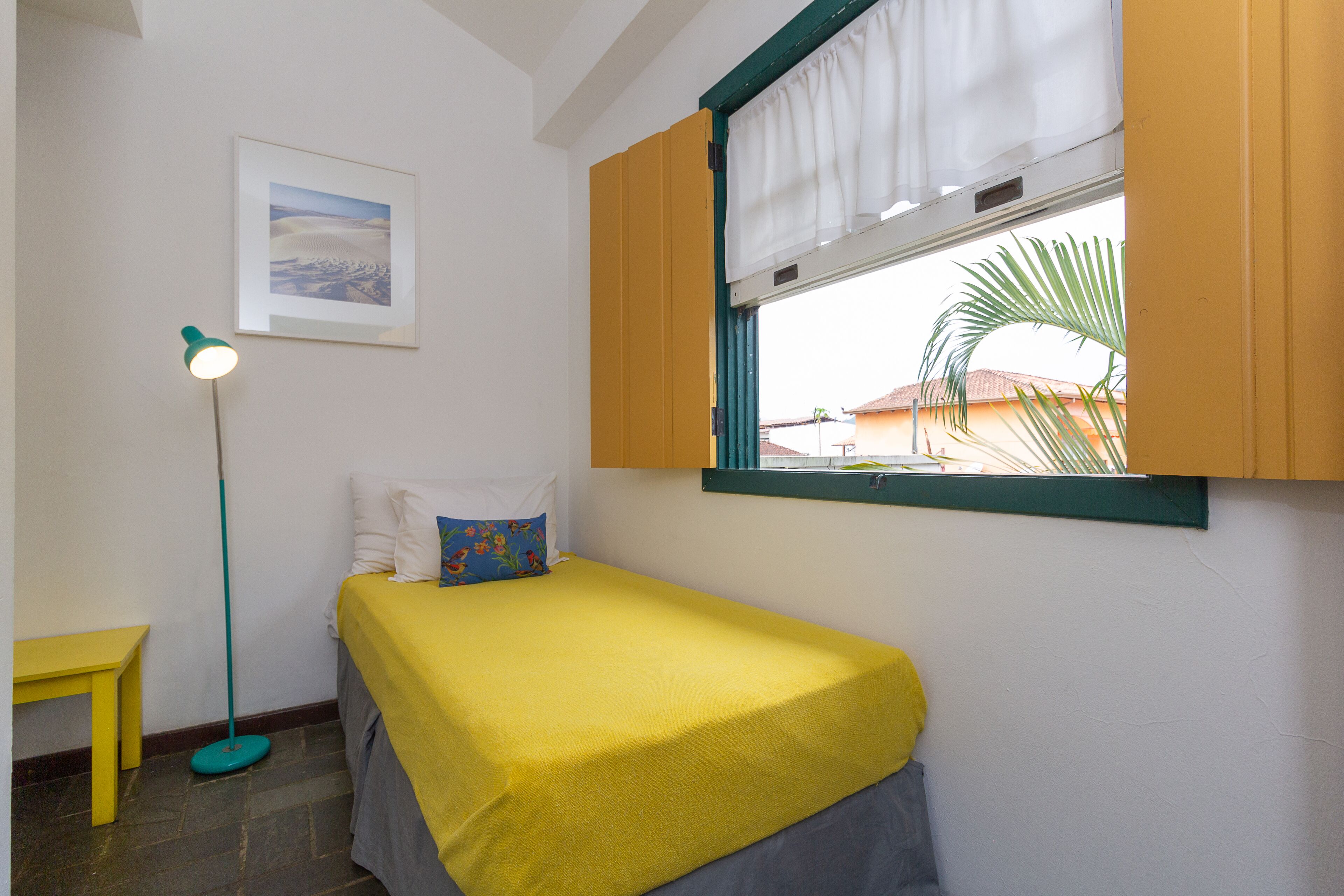 Een kleine, netjes ingerichte kamer met een bed, een bureau, een vloerlamp en een raam met uitzicht op palmbomen.