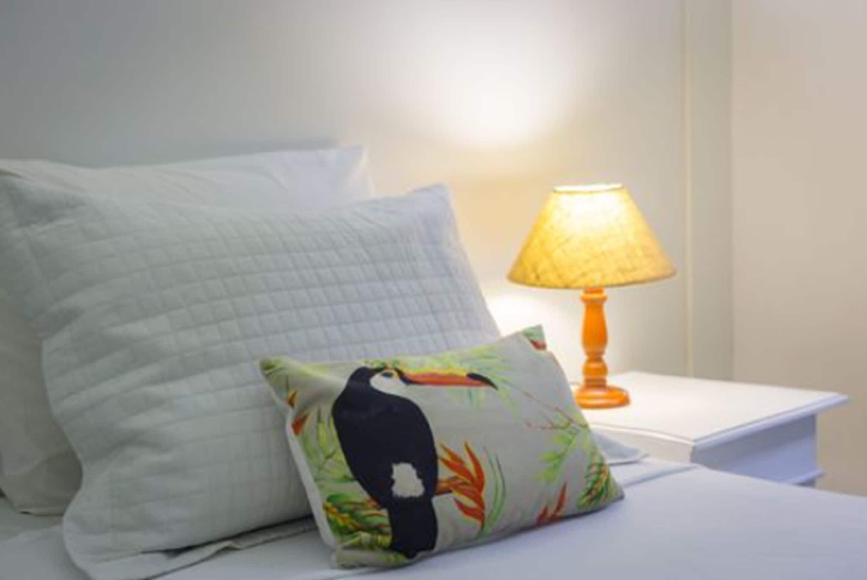 Una cama con ropa de cama blanca, una mesita de noche con lámpara y una almohada con diseño de tucán.