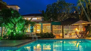 A swimming pool with lounge chairs and a gazebo, surrounded by lush greenery and a building with large windows. - Pousada do Principe (Paraty)