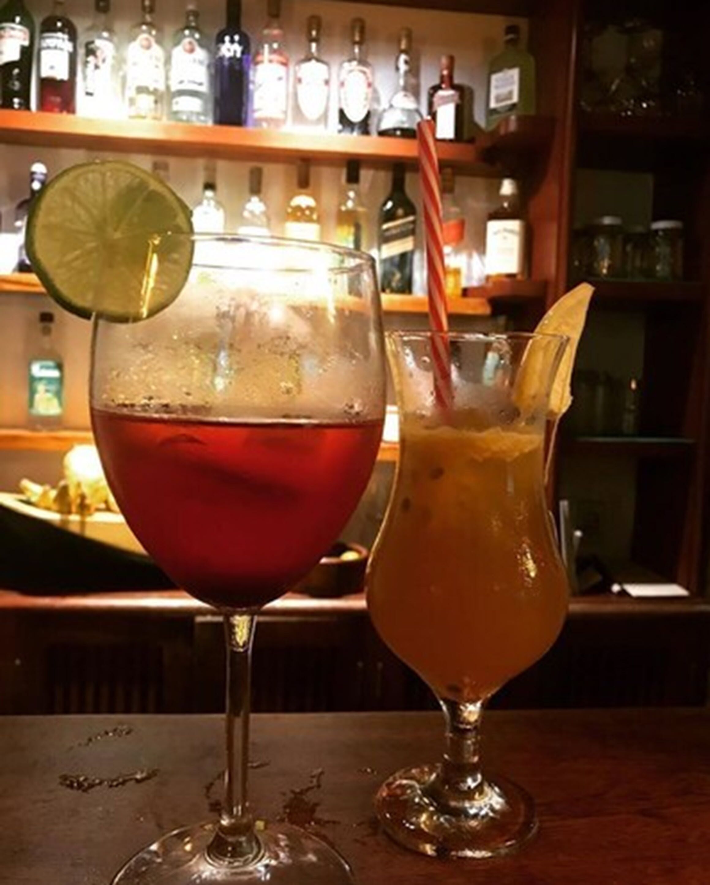 Dos cócteles sobre una barra, con una estantería llena de botellas de licor al fondo.