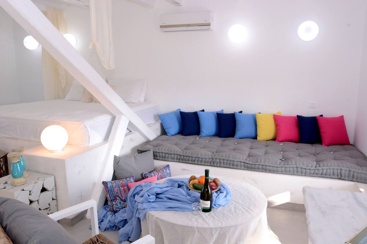 A cozy, well-lit room with a bed, a sofa with colorful cushions, a small table with a bottle and fruit, and a white staircase.