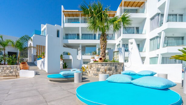 A modern hotel with a rooftop pool area featuring a circular blue lounge with cushions, a palm tree, and a stone wall with potted plants. - Kouros Home Hotel (Rhodes)